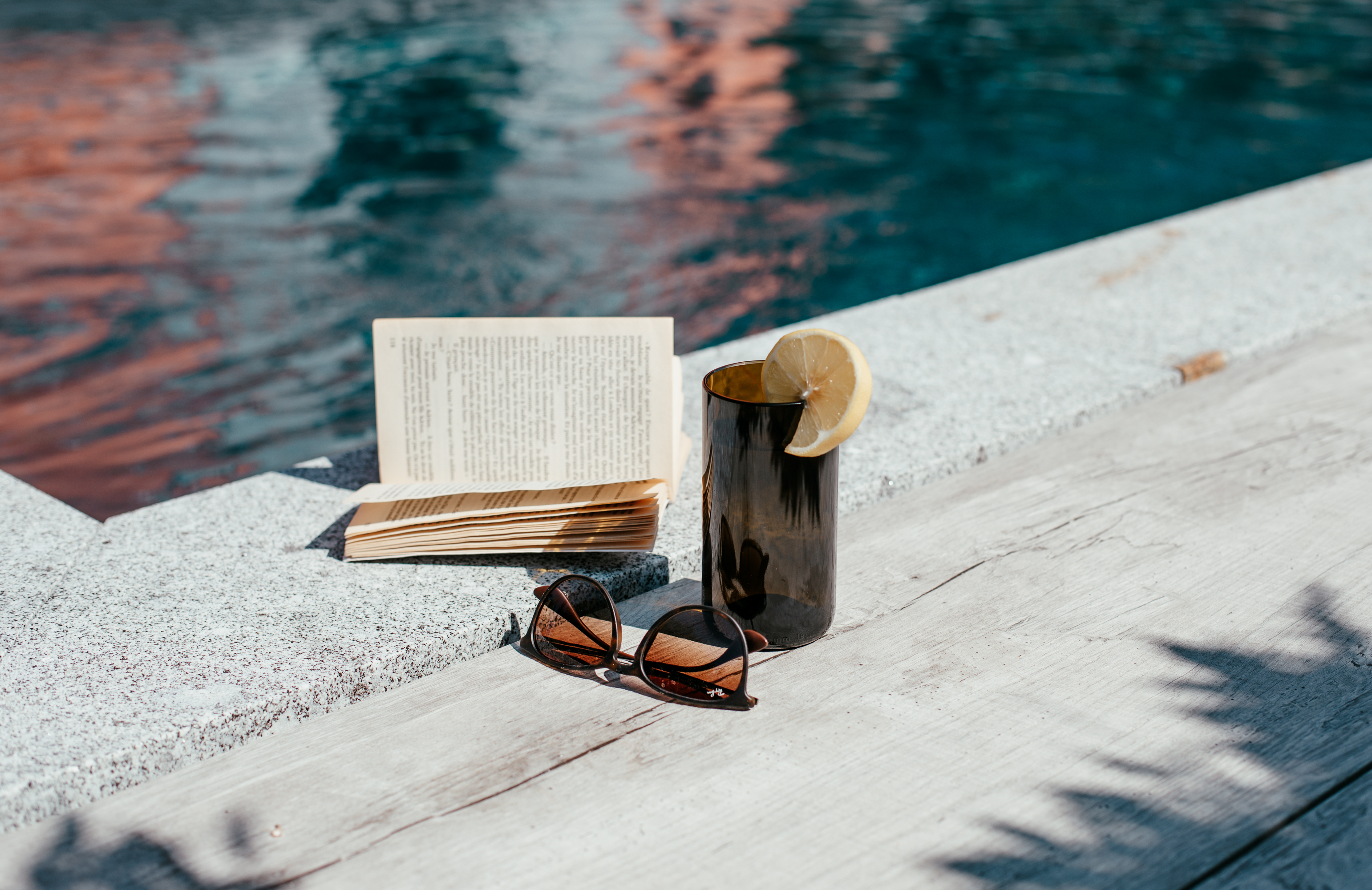Verre brun de la marque kulô posé au bord d'une piscine avec un livre et des lunettes de soleil.